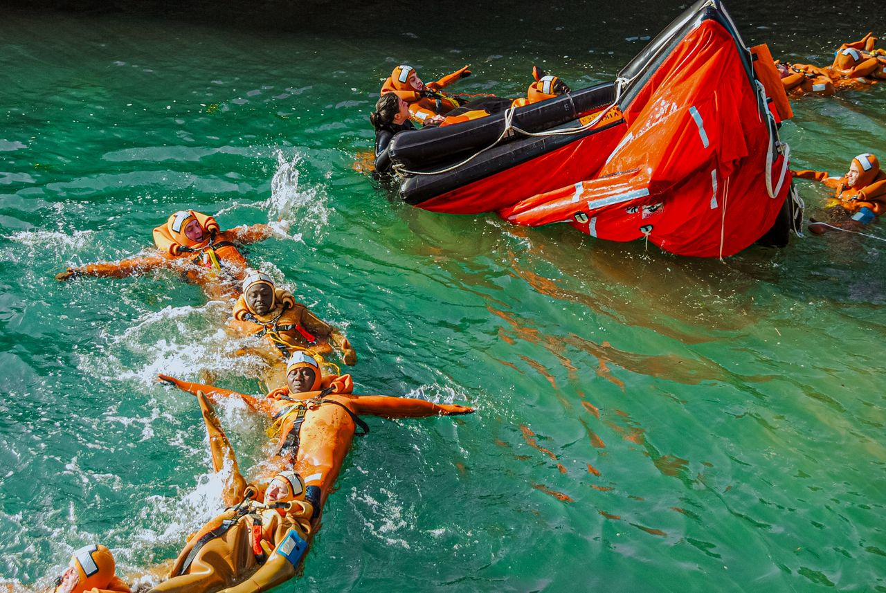 Group of adults practicing lifeboat safety drill in the water wearing life jackets.